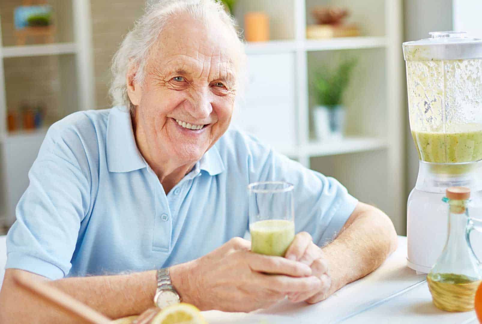 Smiling older man holding a glass of green smoothie while sitting next to the blender that was used to make it
